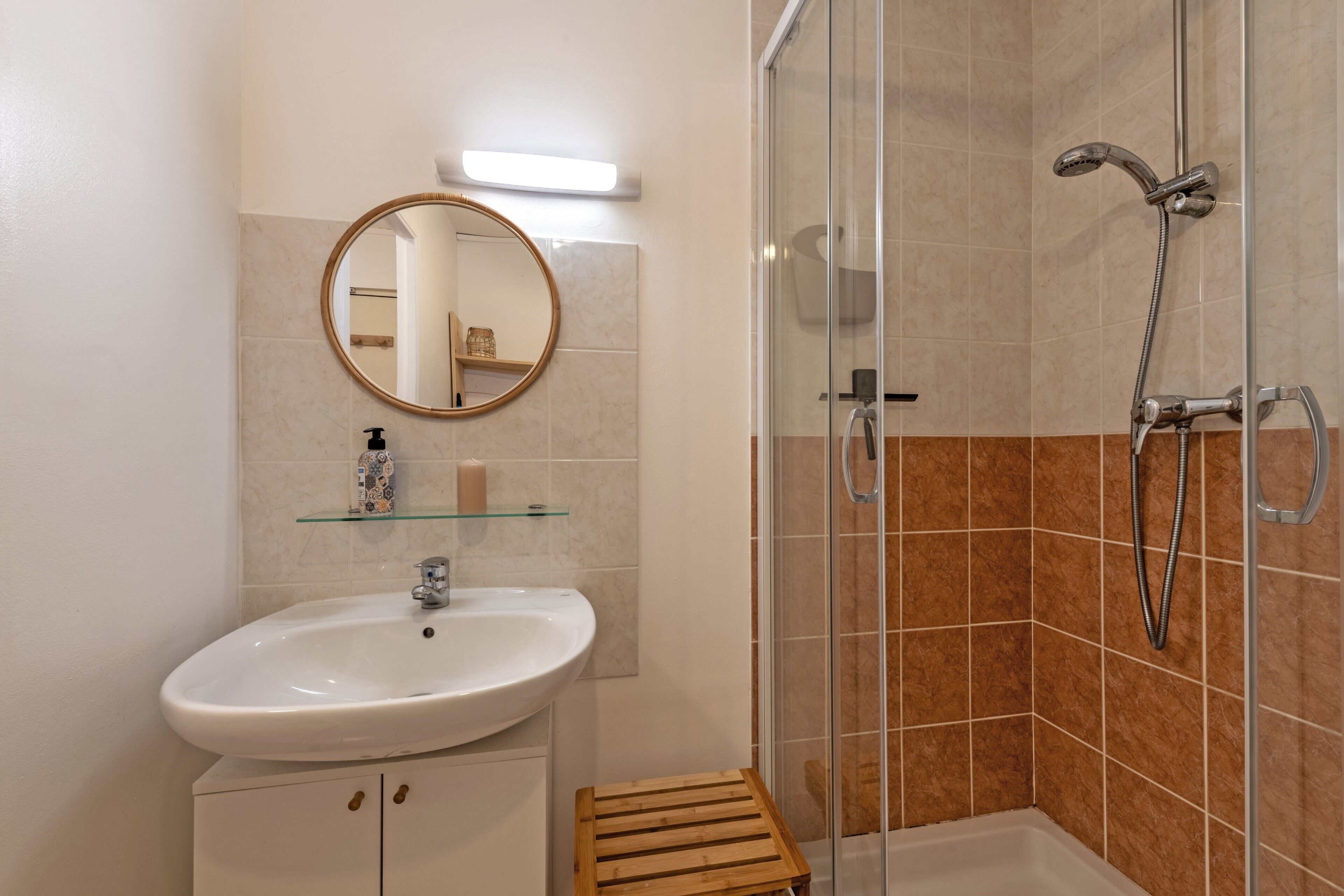 Une salle de bain équipée d’un lavabo, d’un miroir, d’une douche et d’un petit tabouret en bois.