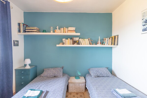 Een slaapkamer met twee enkele bedden, een nachtkastje met een lamp, een boekenkast en een klein mandje.