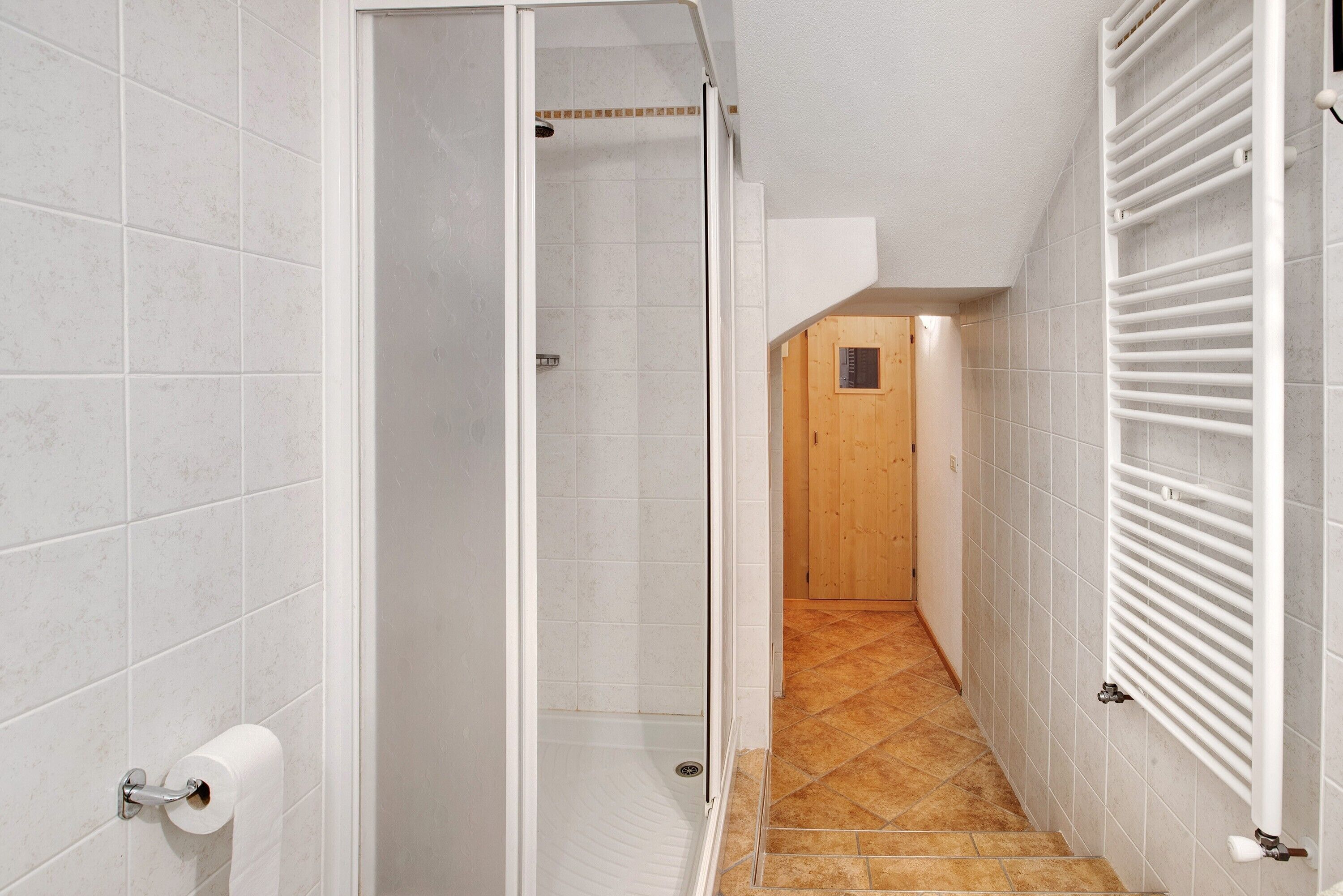 Un couloir étroit, avec une zone douche sur la gauche, une porte en bois au fond et un radiateur blanc sur la droite.