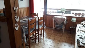 A dining area with a table and chairs, a small table with a lamp, and a view through a window.