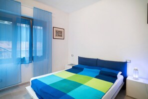 Una camera da letto con un letto, un comodino, una lampada da comodino, una finestra con tende leggere e un quadro appeso al muro.