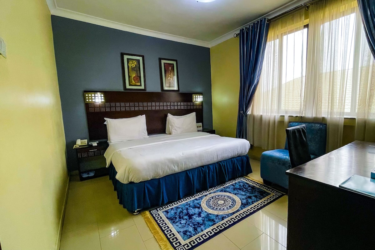 A hotel room with a bed, a desk, a chair, a nightstand, and two wall-mounted lamps.