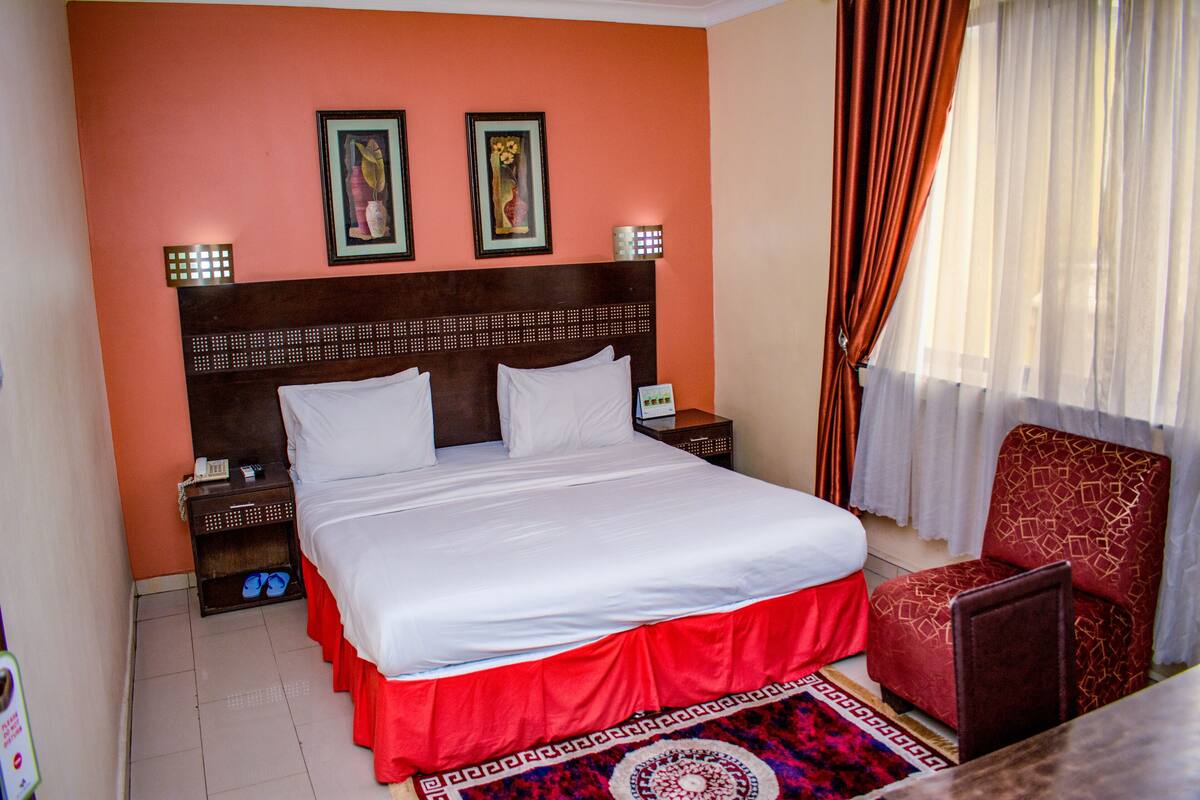 A hotel room with a large bed, a red skirted bed frame, two bedside tables with lamps, a chair, and a window with curtains.