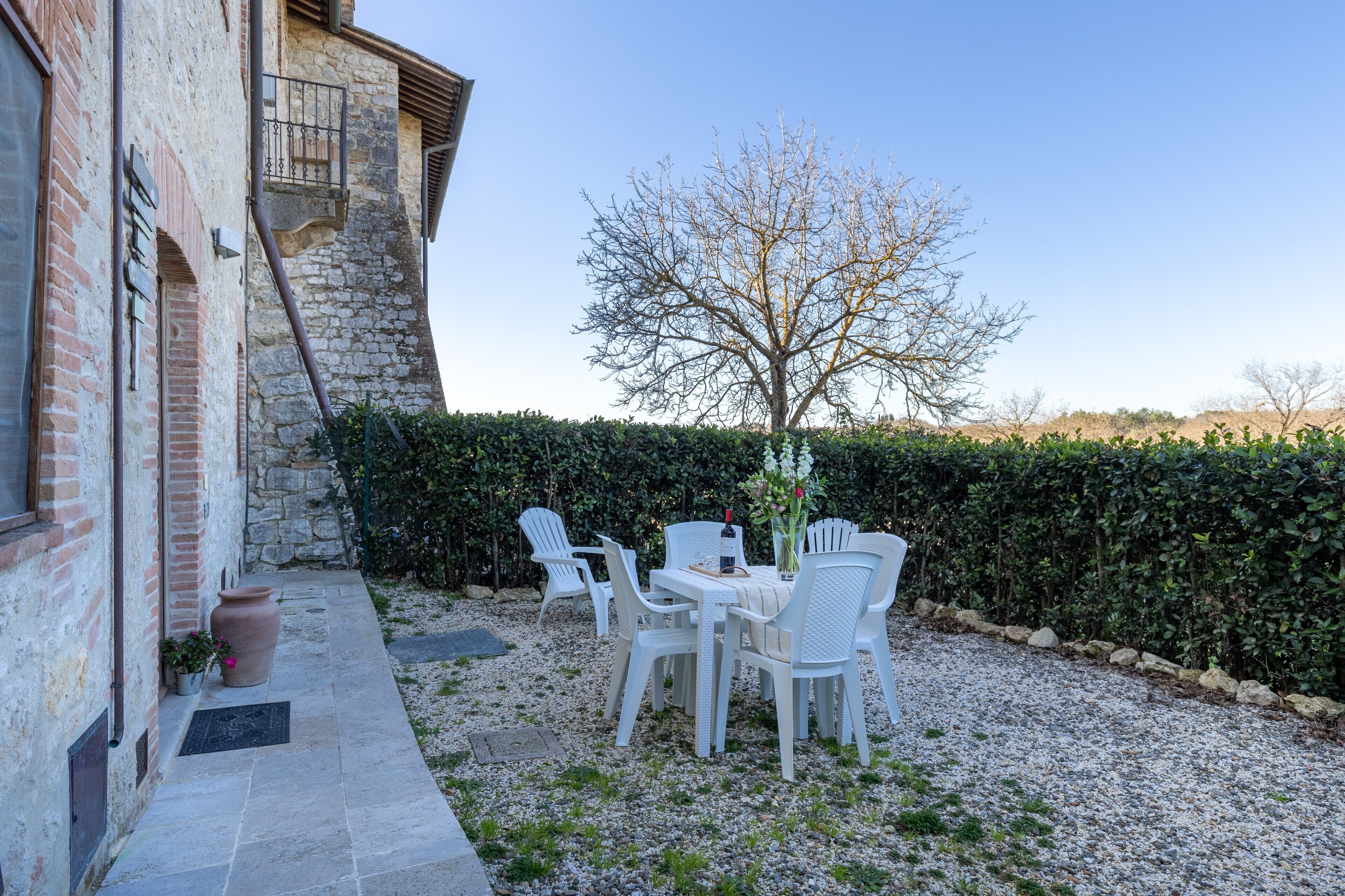 Un patio exterior con sillas y una mesa blancos, un muro de piedra y un árbol.