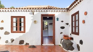 Un edificio blanco con parches de piedra, dos ventanas de madera, un patio con baldosas y una escalera visible que conduce al interior.