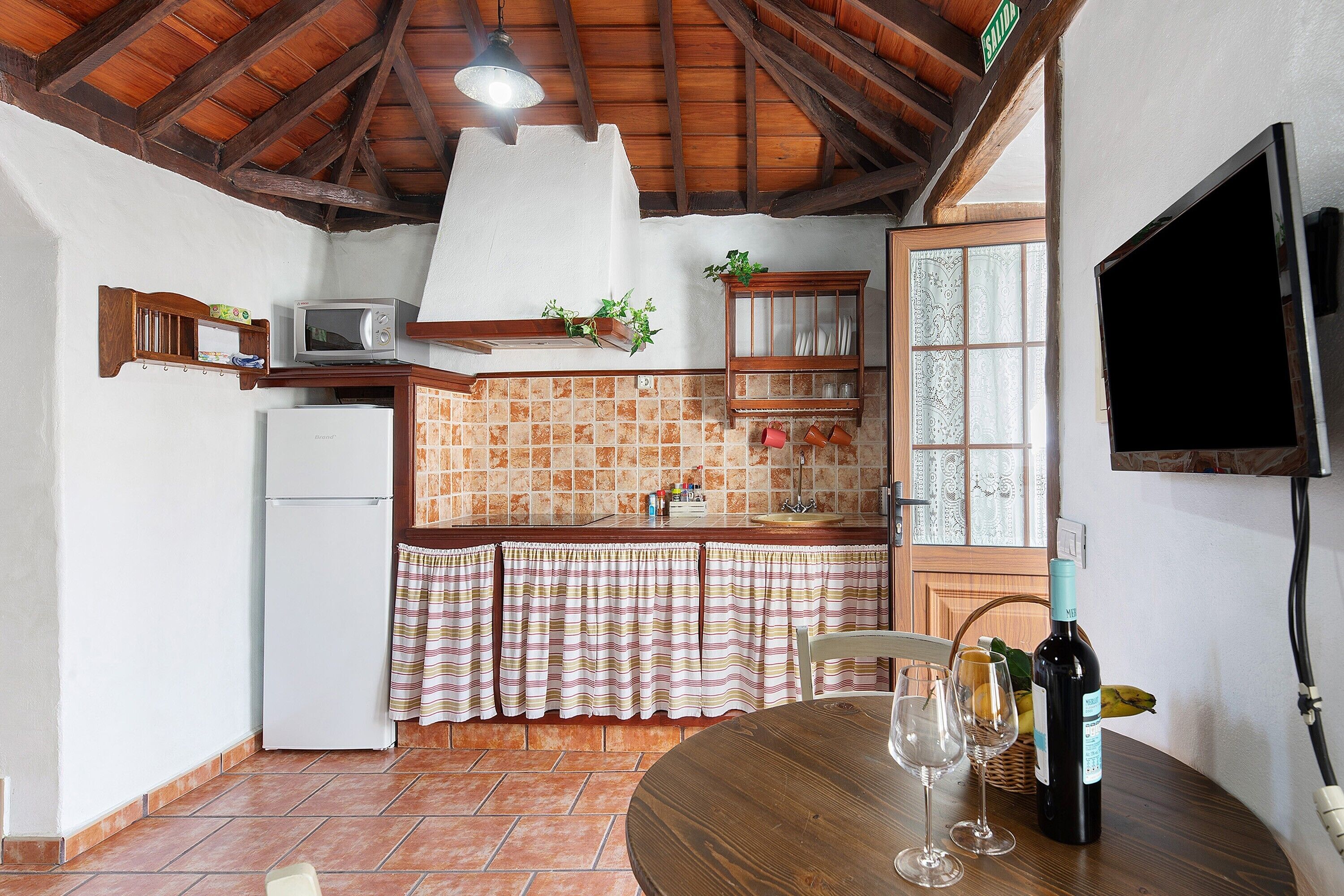 A kitchen with a white refrigerator, a microwave, a wooden cabinet, a window with curtains, a wooden table with wine glasses, and a bottle of wine.