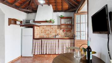 A kitchen with a white refrigerator, a microwave, a wooden cabinet, a window with curtains, a wooden table with wine glasses, and a bottle of wine.