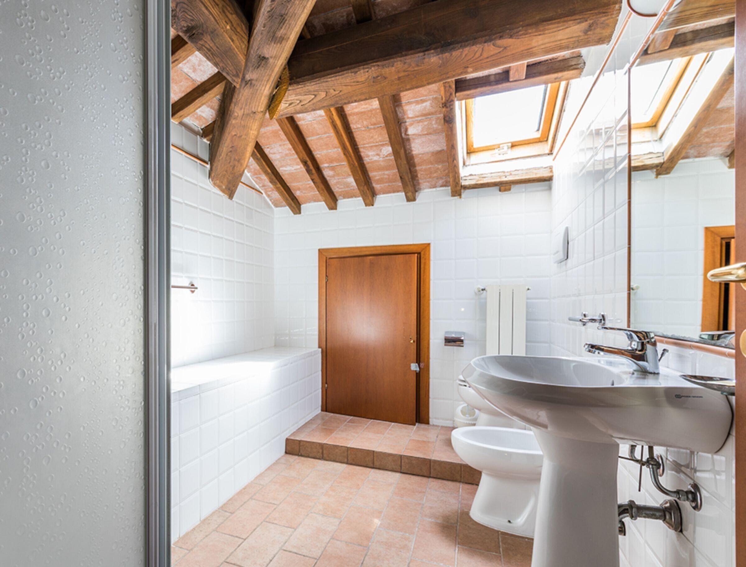 Baño con techo de madera, tragaluz, azulejos blancos, puerta de madera, lavabo blanco y un inodoro.