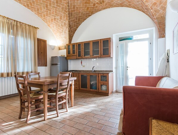 A kitchen with wooden cabinets, a dining table with chairs, a refrigerator, and a red sofa. - Holiday Home 'Podere Simonetta - A6' with Shared Pool, Private Terrace and Wi-Fi (Cecina)