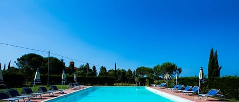 A rectangular swimming pool with a tiled deck, surrounded by lounge chairs and umbrellas, under a clear blue sky.