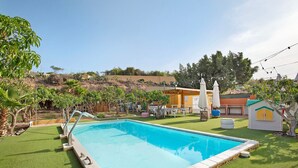 Une piscine entourée d’une terrasse, une petite maison de jeu et un espace clos avec des arbres et des arbustes.