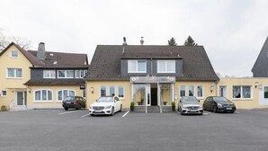 Ein Hotel mit einem zentralen Eingang, einem Parkplatz mit vier Autos und einem gelben Gebäude mit grauem Dach.