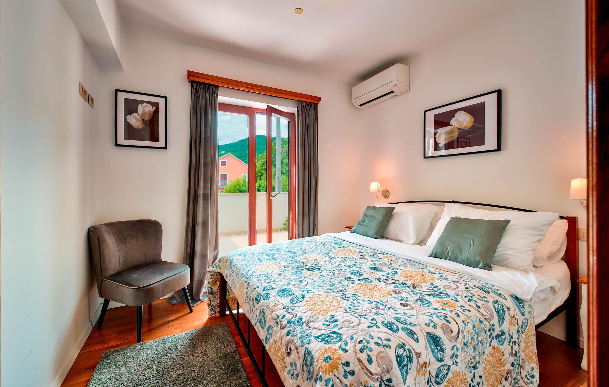 Un dormitorio con cama, una silla, un ventanal con cortinas y dos cuadros en la pared.