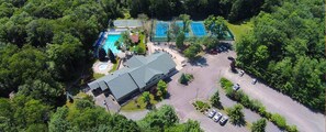 Vista aérea de uma propriedade com piscina, quadras de tênis e estacionamento, tudo cercado por árvores.
