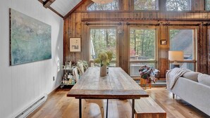 Interior rústico de una cabaña con una mesa de madera, un sofá y grandes ventanas que dan a un bosque.