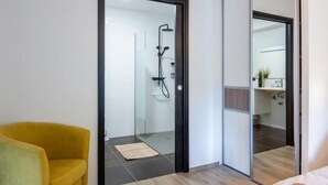 Una habitació d'hotel moderna amb un llit, un otomà groc, una porta de vidre que dona a una zona de banyera i un gran mirall.