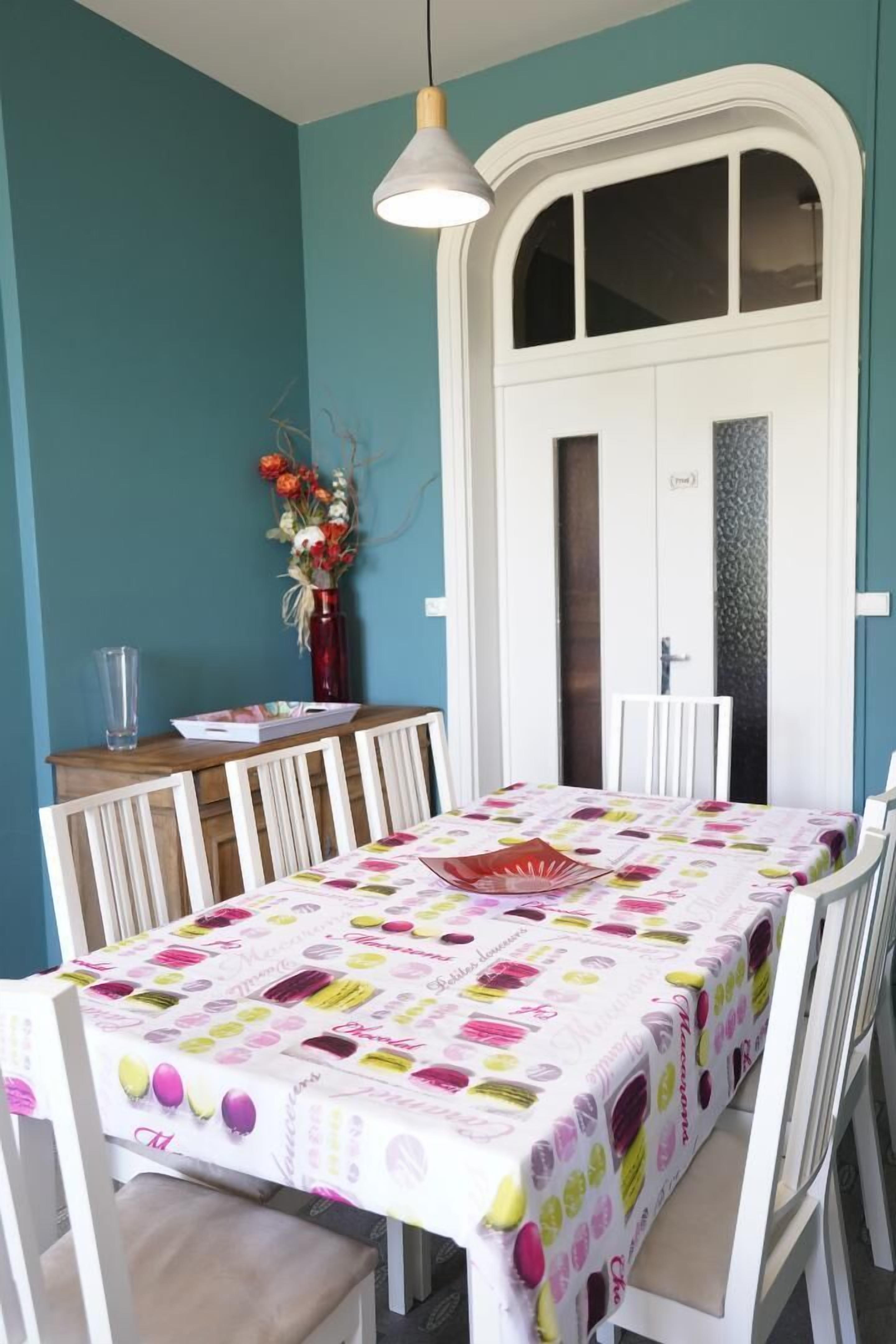 Un comedor con una mesa cubierta con un mantel colorido, sillas blancas, un jarrón con flores y una lámpara colgante.