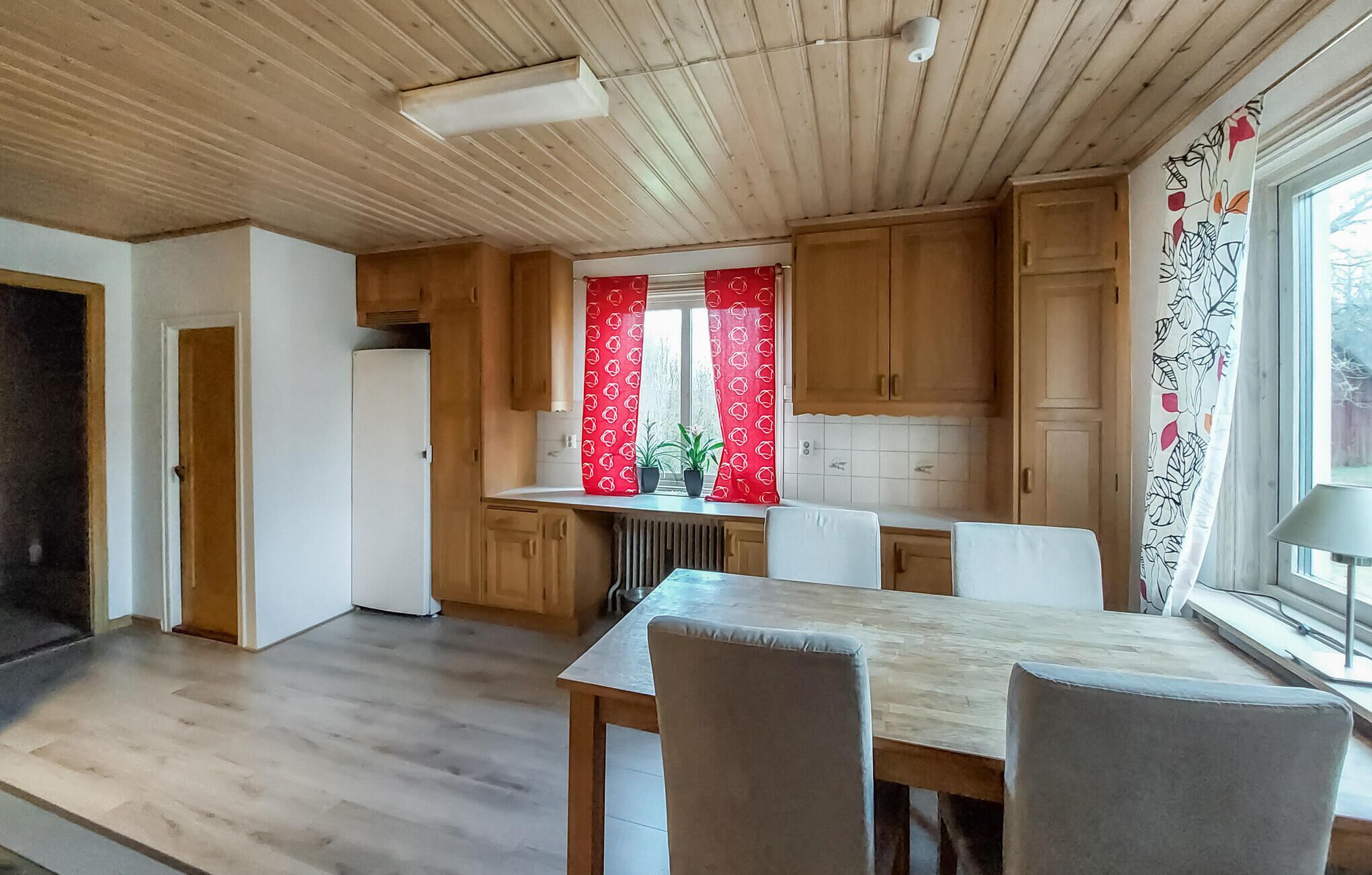 A modern kitchen with wooden cabinets, a dining table with chairs, and a window with red curtains.
