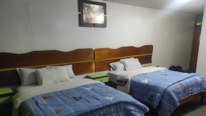 Habitación de hotel con dos camas, cada una con cabecero de madera y ropa de cama azul. Hay una mesita pequeña y una silla, un cuadro en la pared y una maleta en el suelo.
