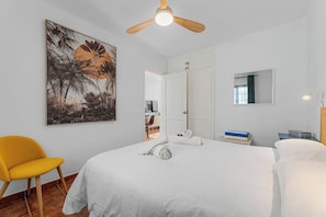 A bedroom with a bed, a ceiling fan, a painting, a chair, and a nightstand with books.