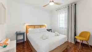 Une chambre d’hôtel avec un lit, un ventilateur de plafond, une table de chevet, une chaise et une fenêtre avec des rideaux.