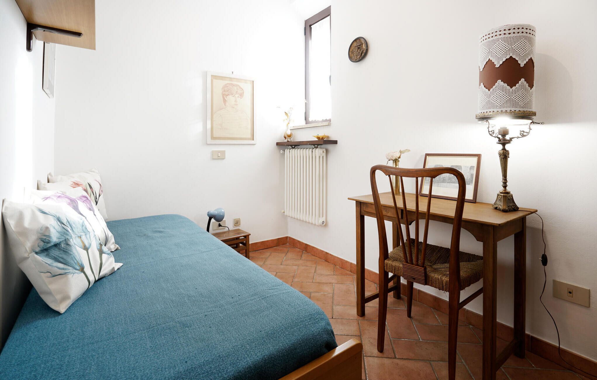 A bedroom with a bed, a desk with a chair, a radiator, and a wall-mounted lamp.