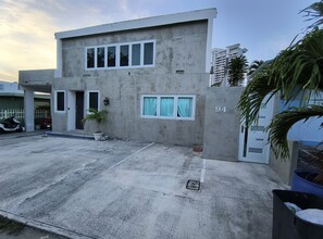 Un bâtiment moderne à deux étages, doté d’une façade en béton, portant le numéro 94, et agrémenté d’un petit jardin devant, d’une plante en pot et d’une seule fenêtre avec des rideaux.