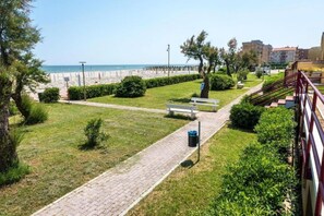 Un sendero pavimentado que lleva a un banco, una zona con césped y vistas a la playa.