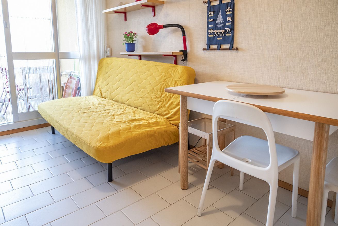 A room with a yellow sofa, a white chair, a wooden table, a red lamp, and a blue wall hanging.