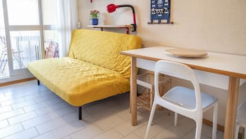 A room with a yellow sofa, a white chair, a wooden table, a red lamp, and a blue wall hanging.