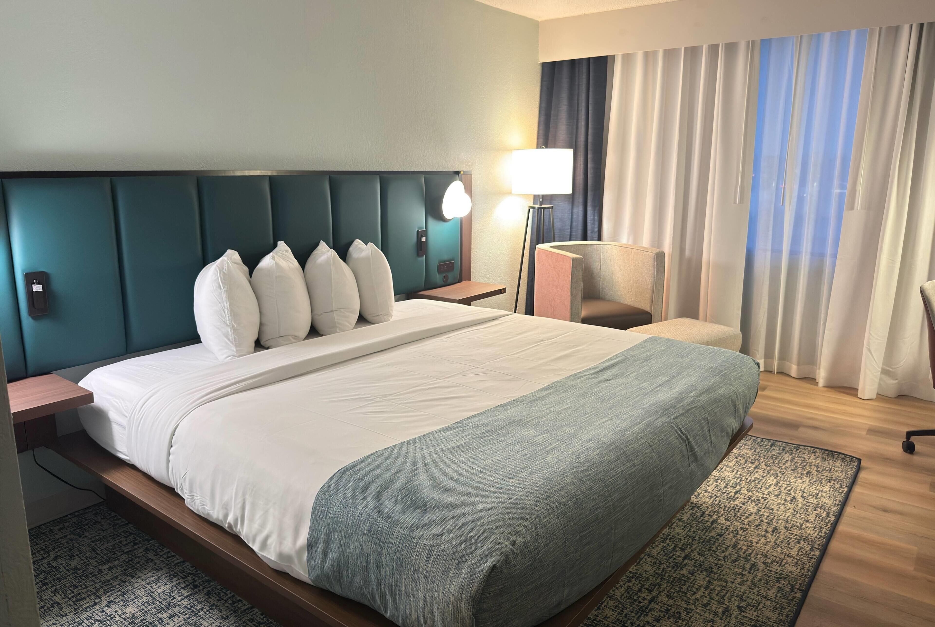 Ein Hotelzimmer mit einem großen Bett, einem Nachttisch, einer Lampe, einem Sessel und Blick ins Freie.