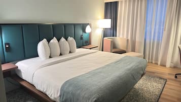 Ein Hotelzimmer mit einem großen Bett, einem Nachttisch, einer Lampe, einem Sessel und Blick ins Freie.