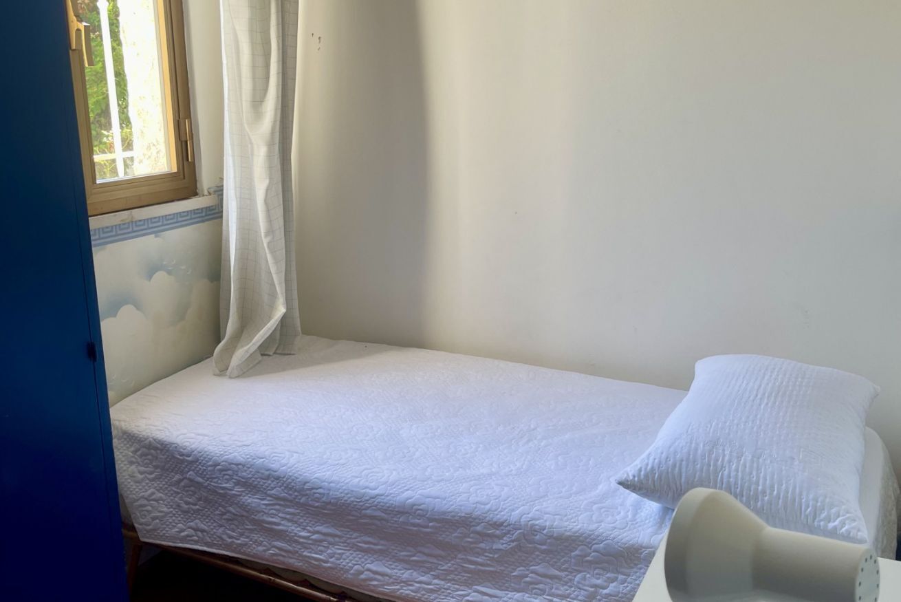 A small, single-bed room with a window, a curtain, and a white pillow.