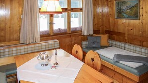 A wooden cabin interior with a dining table, bench seating, a sofa with cushions, a window with curtains, and a painting on the wall. - Ferienhaus - Wald am Arlberg (Wald am Arlberg)