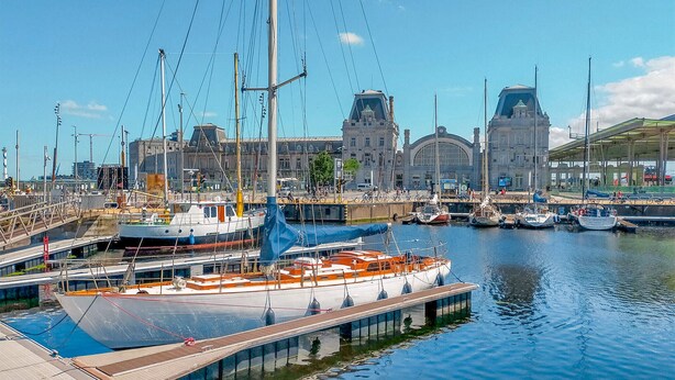 Een jachthaven met aangemeerde boten, een groot gebouw met een klokkentoren, en een overdekte gang.