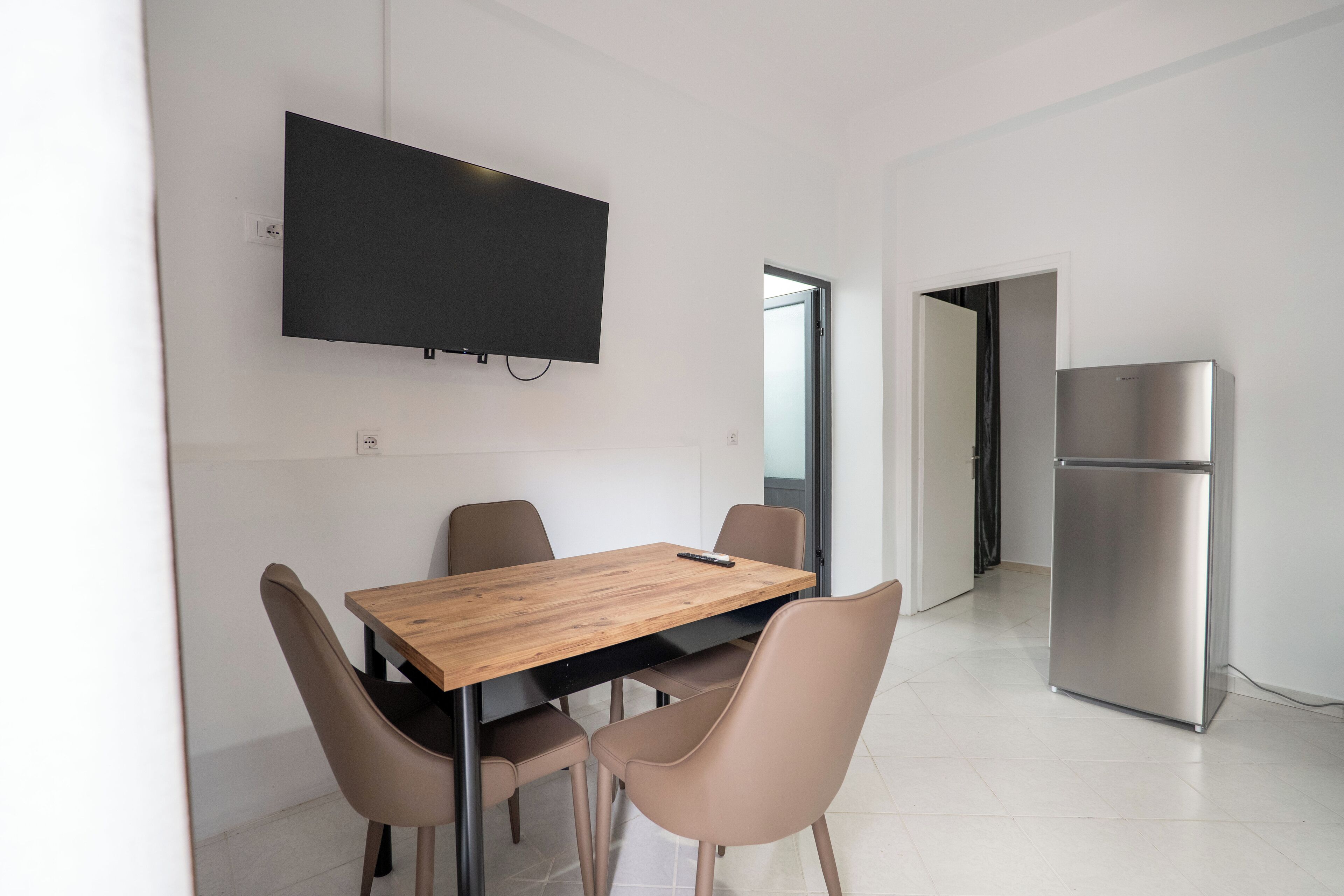 A modern dining area with a wooden table and four chairs, a flat-screen TV mounted on the wall, and a stainless steel refrigerator in the background.