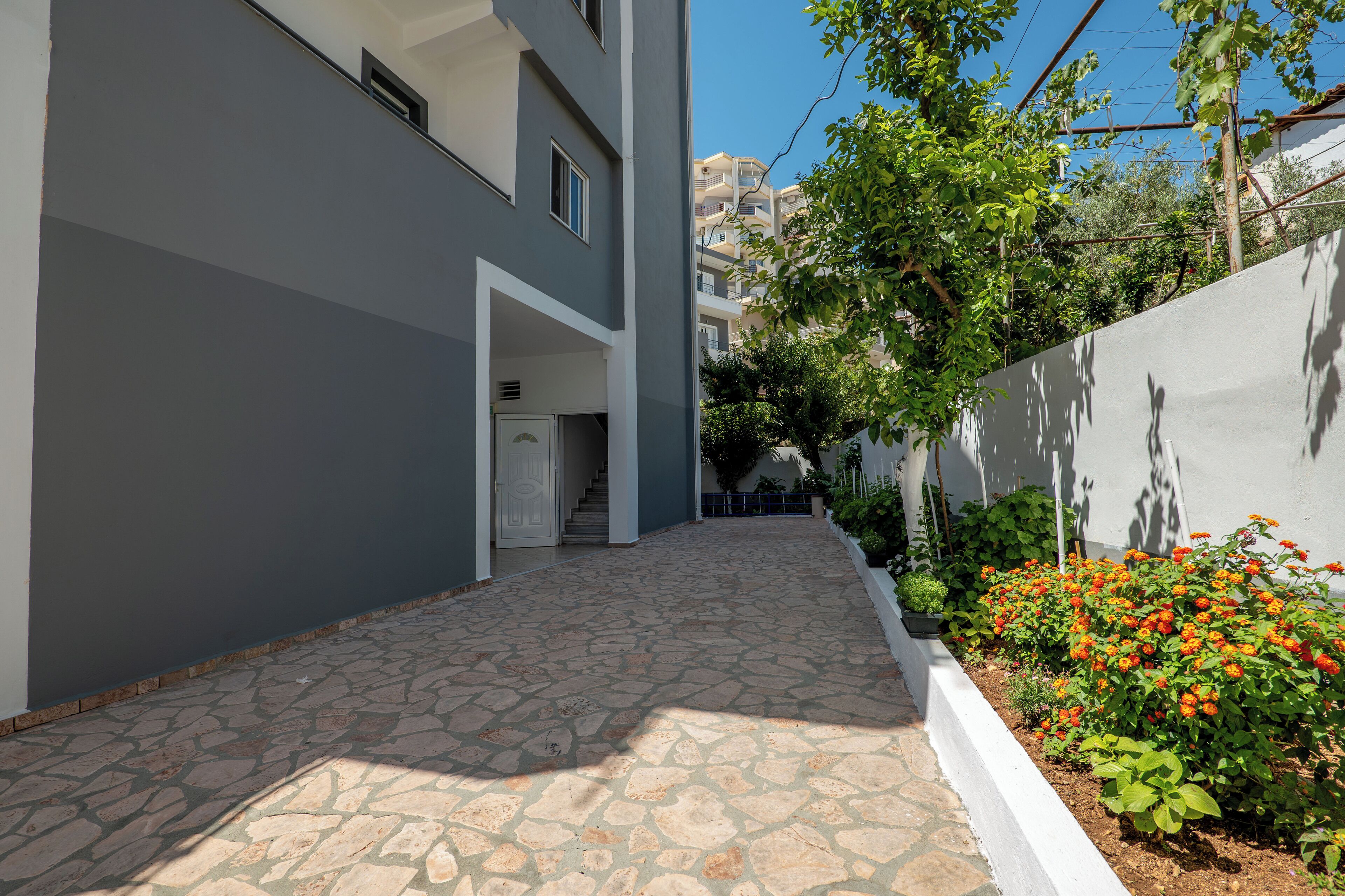 A narrow stone-paved pathway leading to a building entrance, surrounded by greenery and a white wall.
