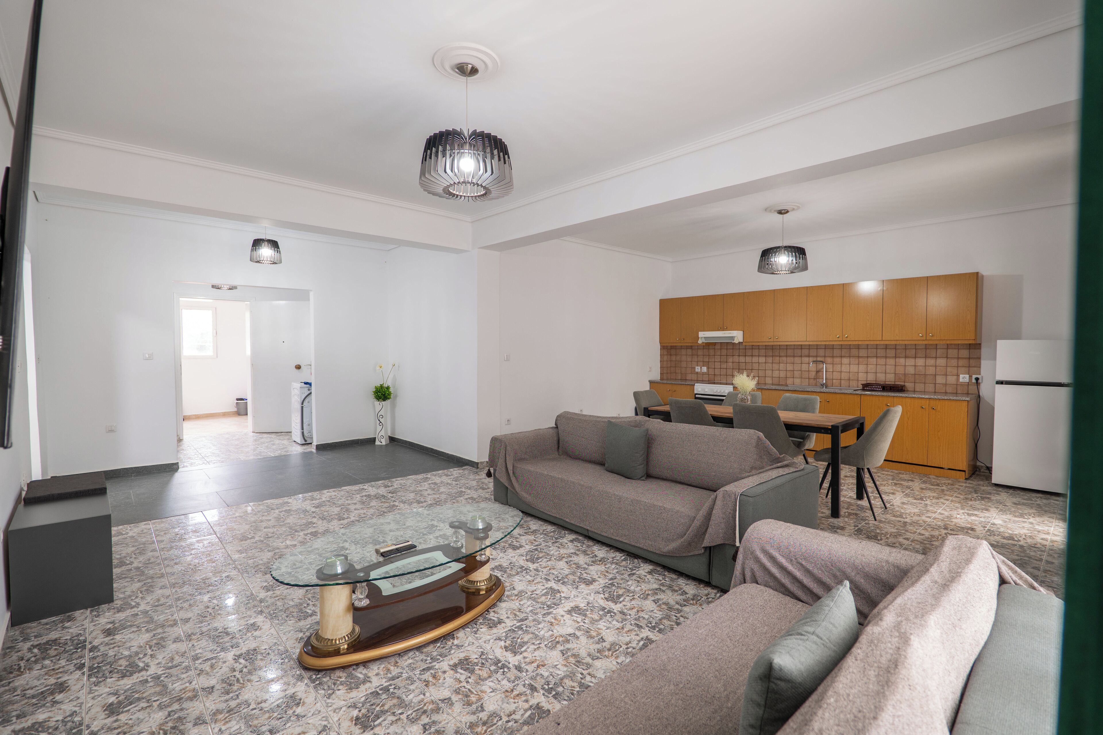 A modern living room with a glass coffee table, a grey sofa, and a kitchen area with wooden cabinets and a tiled backsplash.