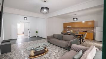 A modern living room with a glass coffee table, a grey sofa, and a kitchen area with wooden cabinets and a tiled backsplash.