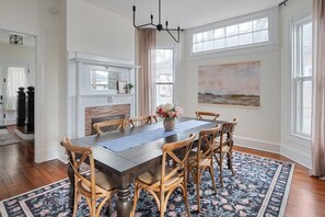 Une salle à manger avec une grande table en bois, six chaises en bois, un arrangement floral en guise de décoration de table, une cheminée et une grande fenêtre offrant une vue dégagée.