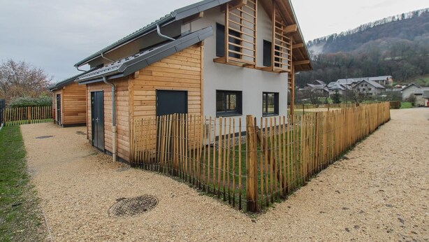 Un bâtiment en bois à deux étages, doté d’un toit en pente, d’une zone délimitée par une clôture et d’une allée gravillonnée.