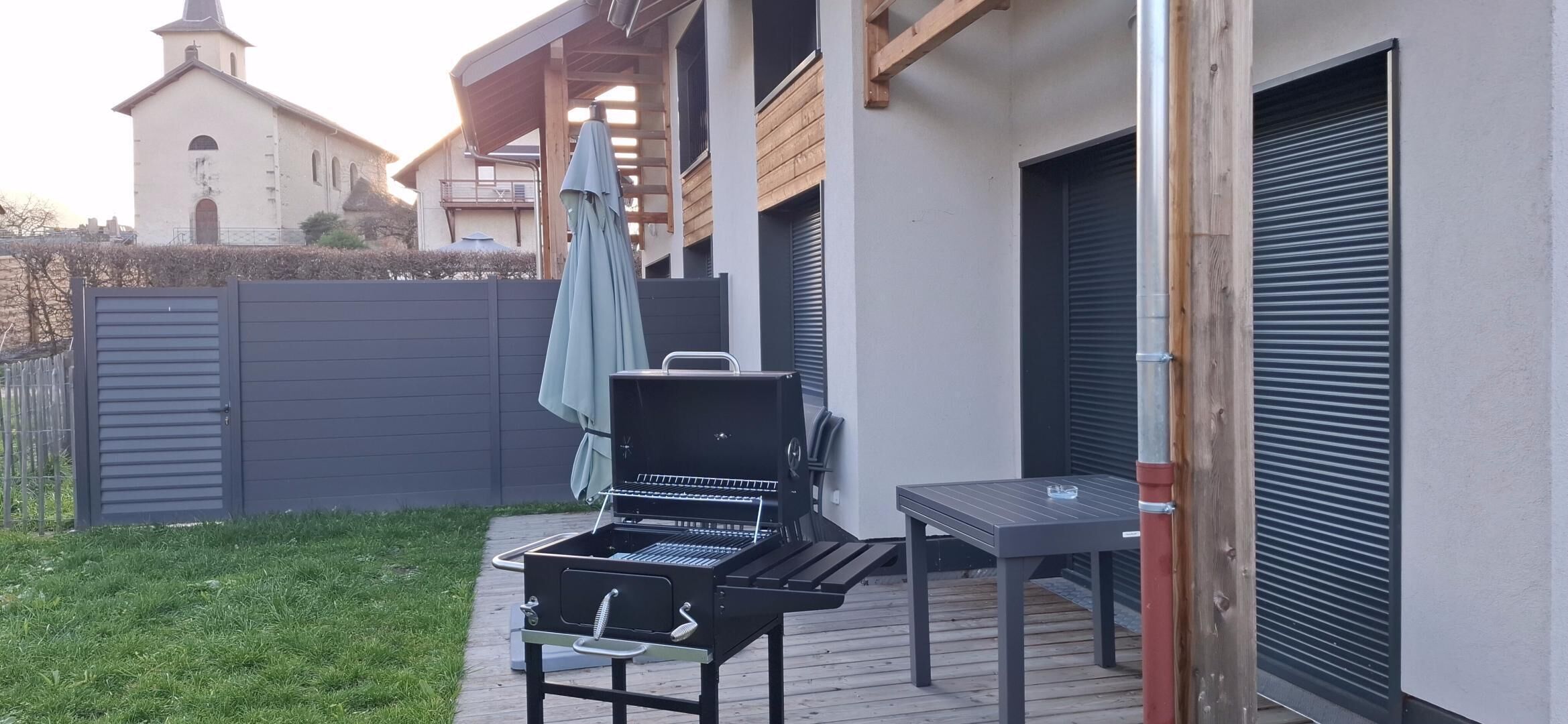 A barbecue grill and table on a wooden deck, with a house and a church in the background.