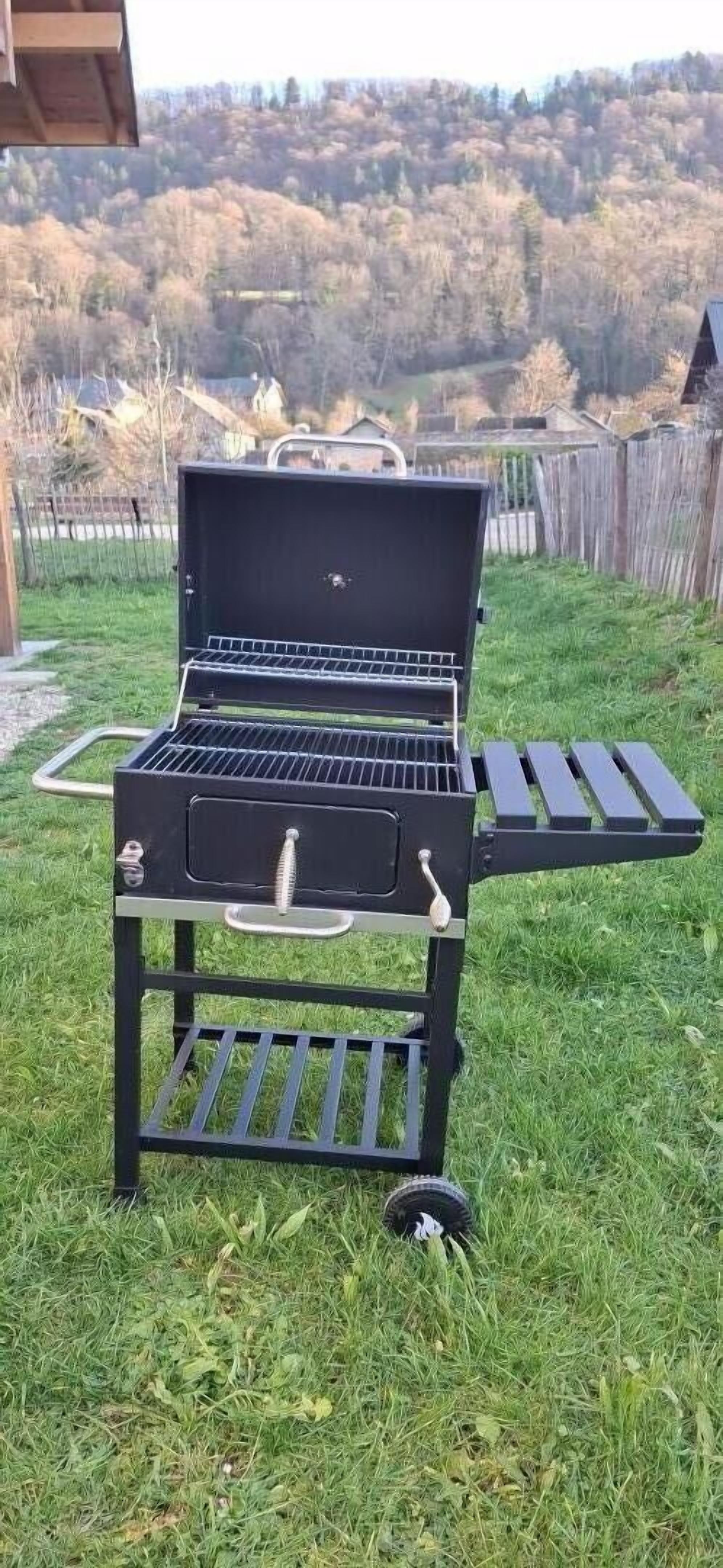 Een draagbare grill met een opklapbare tafel, op een gazon met bebossing op de achtergrond.