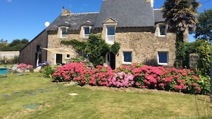 Una casa de piedra con techo inclinado, balcón y jardín lleno de flores rosas.