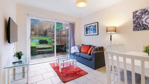 A modern living room with a glass coffee table, a sofa with cushions, a TV mounted on the wall, and a dining area with a white table and chairs. - HP13 3BR Home w/Garden, WiFi &Easy Transport Links (High Wycombe)