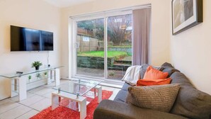 A modern living room with a glass coffee table, a grey sofa with orange cushions, a flat-screen TV mounted on the wall, and a sliding glass door leading to a garden. - HP13 3BR Home w/Garden, WiFi &Easy Transport Links (High Wycombe)