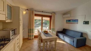A compact living space with a kitchenette, a dining area with a table and chairs, a sofa, and a balcony with a view of greenery. - Holiday apartment mit Klimaanlage und Meerblick (Badesi)