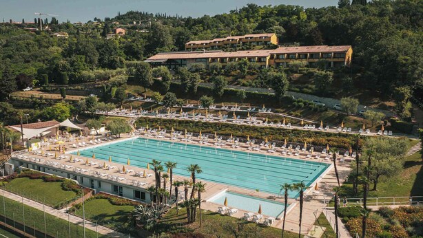 A large swimming pool with multiple lanes, surrounded by sun loungers and umbrellas, set against a backdrop of lush greenery and buildings.