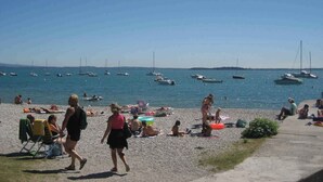 A pebbled beach with people sunbathing and swimming. Boats are anchored in the water.