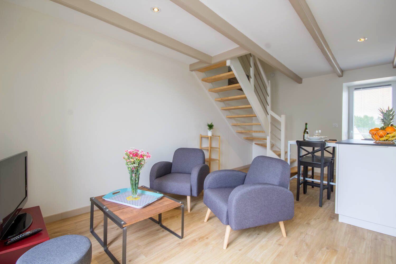 Un salon moderne avec un escalier, des fauteuils gris, une petite table sur laquelle est posé un vase de fleurs, et un coin cuisine avec un panier de fruits.